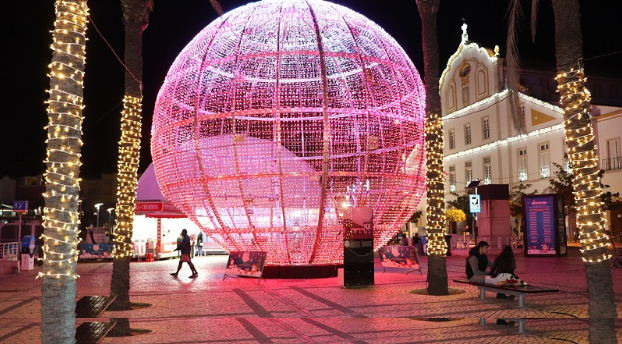 Grande esfera iluminada em tons de rosa e branco, decorando uma praça no centro da cidade, rodeada por palmeiras com luzes douradas e um edifício histórico ao fundo, criando um cenário natalício vibrante e acolhedor.