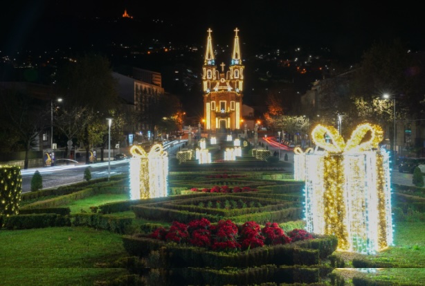 Jardim decorado com enormes presentes iluminados e flores vermelhas, tendo ao fundo uma igreja com iluminação dourada, criando um cenário natalício encantador durante a noite.