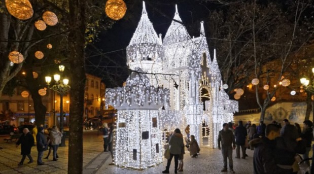 strutura iluminada em forma de castelo no centro de Viseu, decorada com luzes brancas brilhantes, rodeada por árvores com decorações luminosas e pessoas a desfrutar do ambiente festivo.