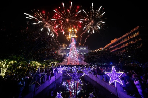 Grande árvore de Natal 3D iluminada na Praça do General Humberto Delgado, no Porto, rodeada por decorações de estrelas luminosas e com fogos de artifício ao fundo, criando um ambiente mágico e festivo.