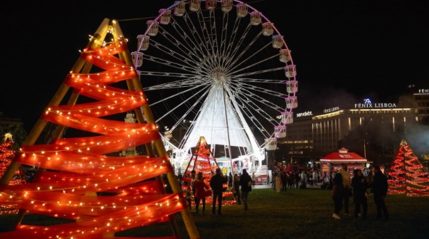 Ambiente natalício no Wonderland Lisboa, com uma roda gigante iluminada ao fundo, árvores de Natal decoradas com luzes vermelhas e douradas, e várias pessoas a passear pelo local, no Parque Eduardo VII.