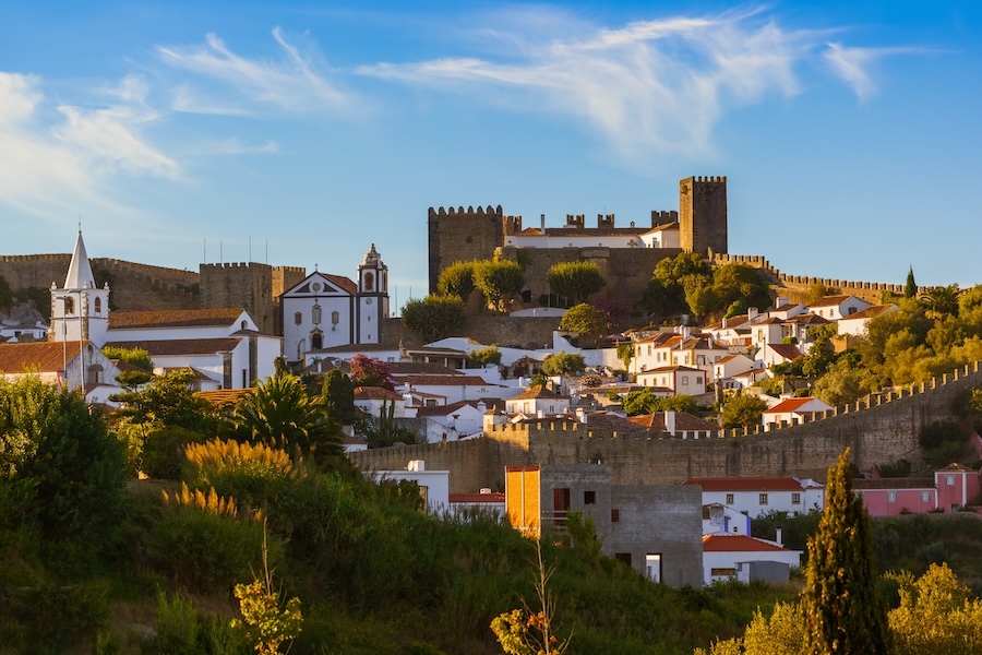 Vista panorâmica da vila medieval de Óbidos, em Portugal, com as suas muralhas imponentes, o castelo histórico no topo e as casas tradicionais de telhados vermelhos espalhadas pela paisagem, destacando-se contra um céu azul e colinas verdes.