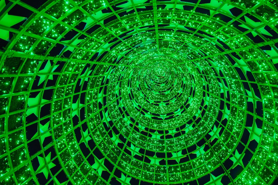 Perspetiva interior de uma estrutura iluminada em forma de túnel, com milhares de luzes verdes brilhantes e padrões de estrelas, criando um efeito visual hipnotizante e festivo, típico de decorações natalícias.